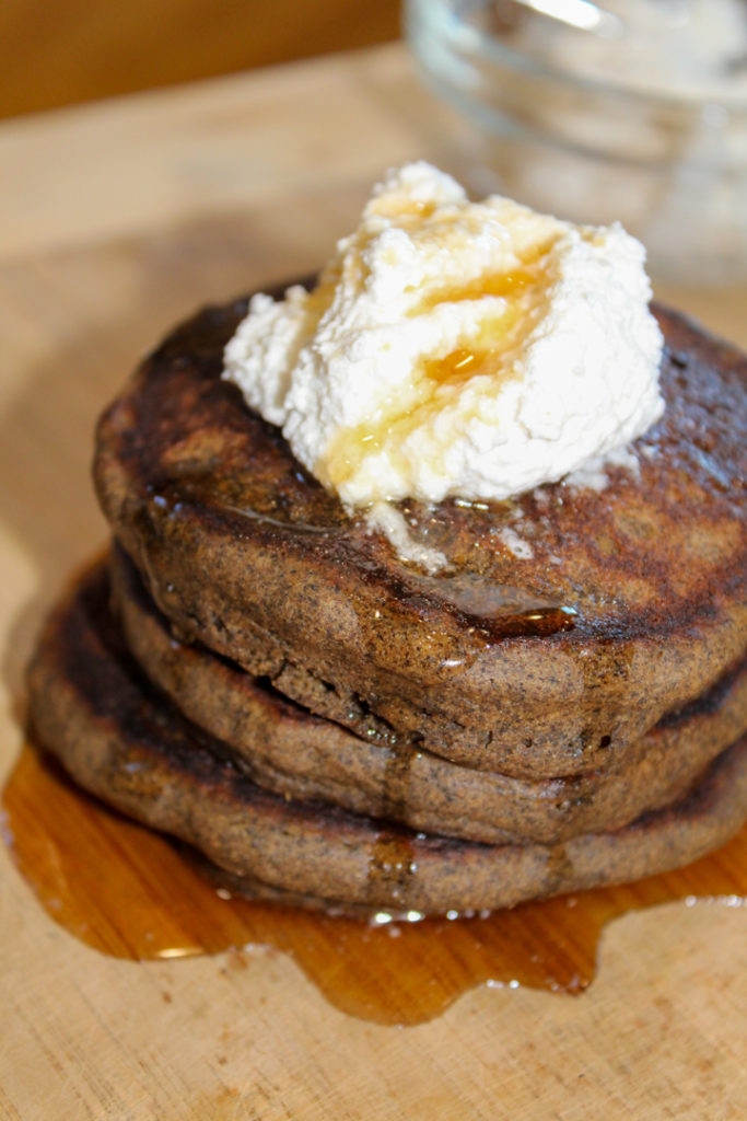 Stack of three pumpkin spice pancakes sitting on a wooden cutting board topped with dairy free whipped topping and maple syrup