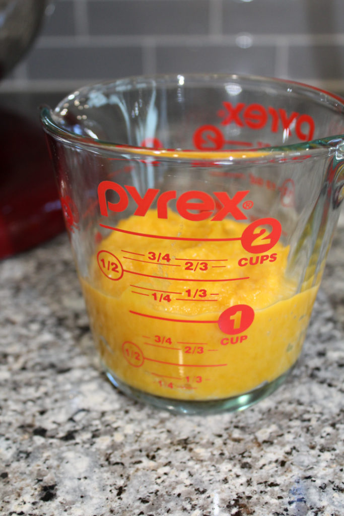 1 cup of pumpkin puree in a glass 2 cup measuring cup. The measuring cup is sitting on the counter in front of a red kitchen aid mixer.