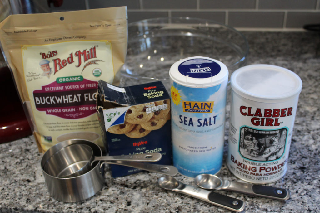 Dry ingredients for pumpkin spice pancakes on the counter with a glass bowl behind them and measuring cups and spoons in front of them.