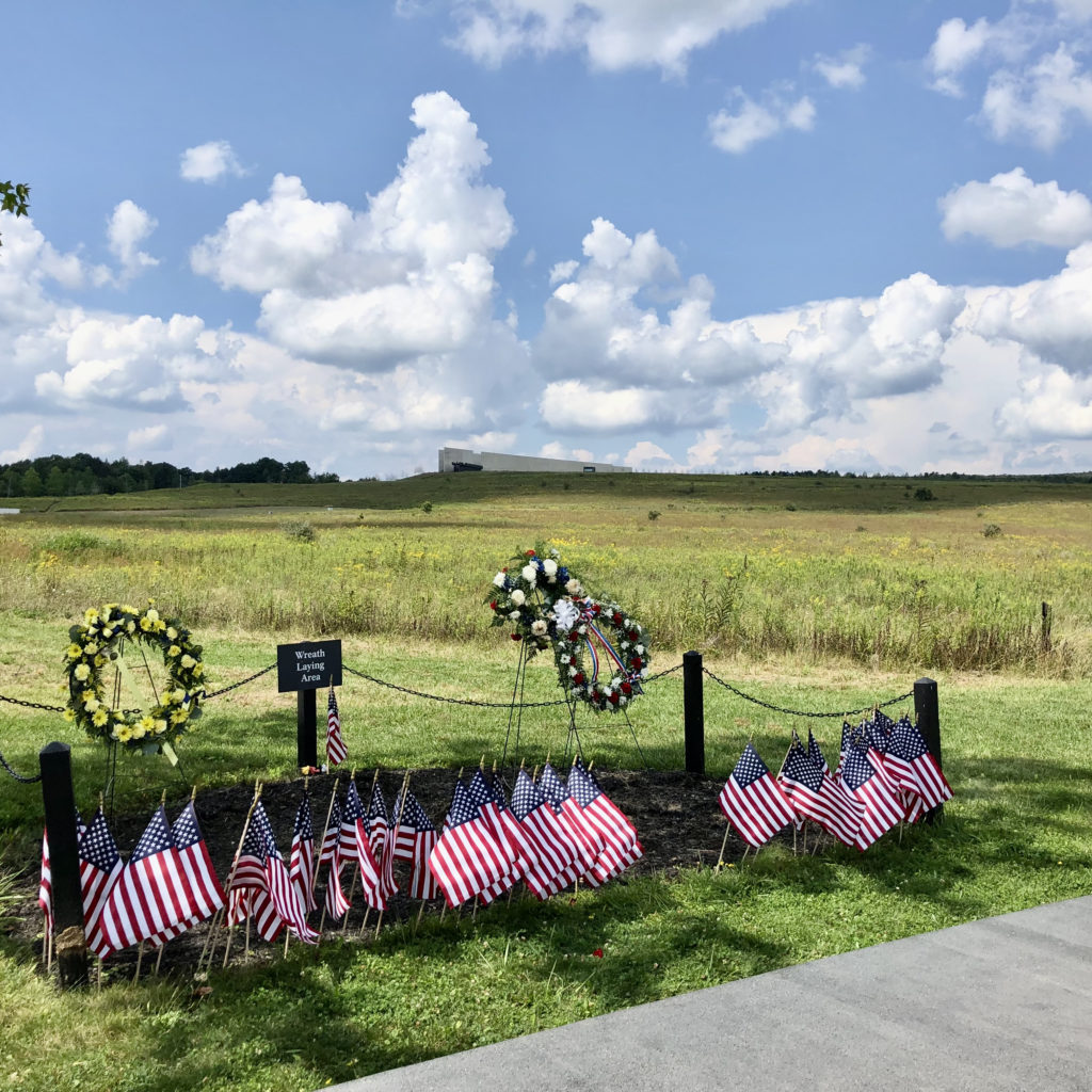 flags and wreaths in the grass next to a sidewalk at the 93 memorial field. behind the wreaths are a few posts, rope, and a sign.