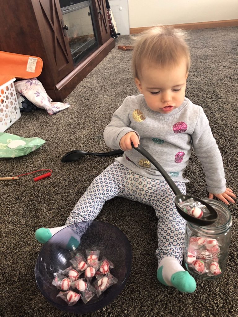 Toddler using eye movement skills to track the spoon she is holding as she scoops and dumps peppermints from a bowl into a mason jar. The bowl is between her legs and the jar is to her left. She is using her right hand to move the spoon.